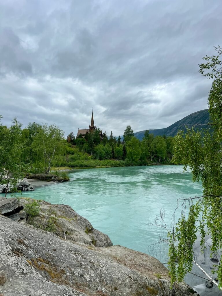 Historic Lom Stave Church overlooking a turquoise glacial river in Norway.